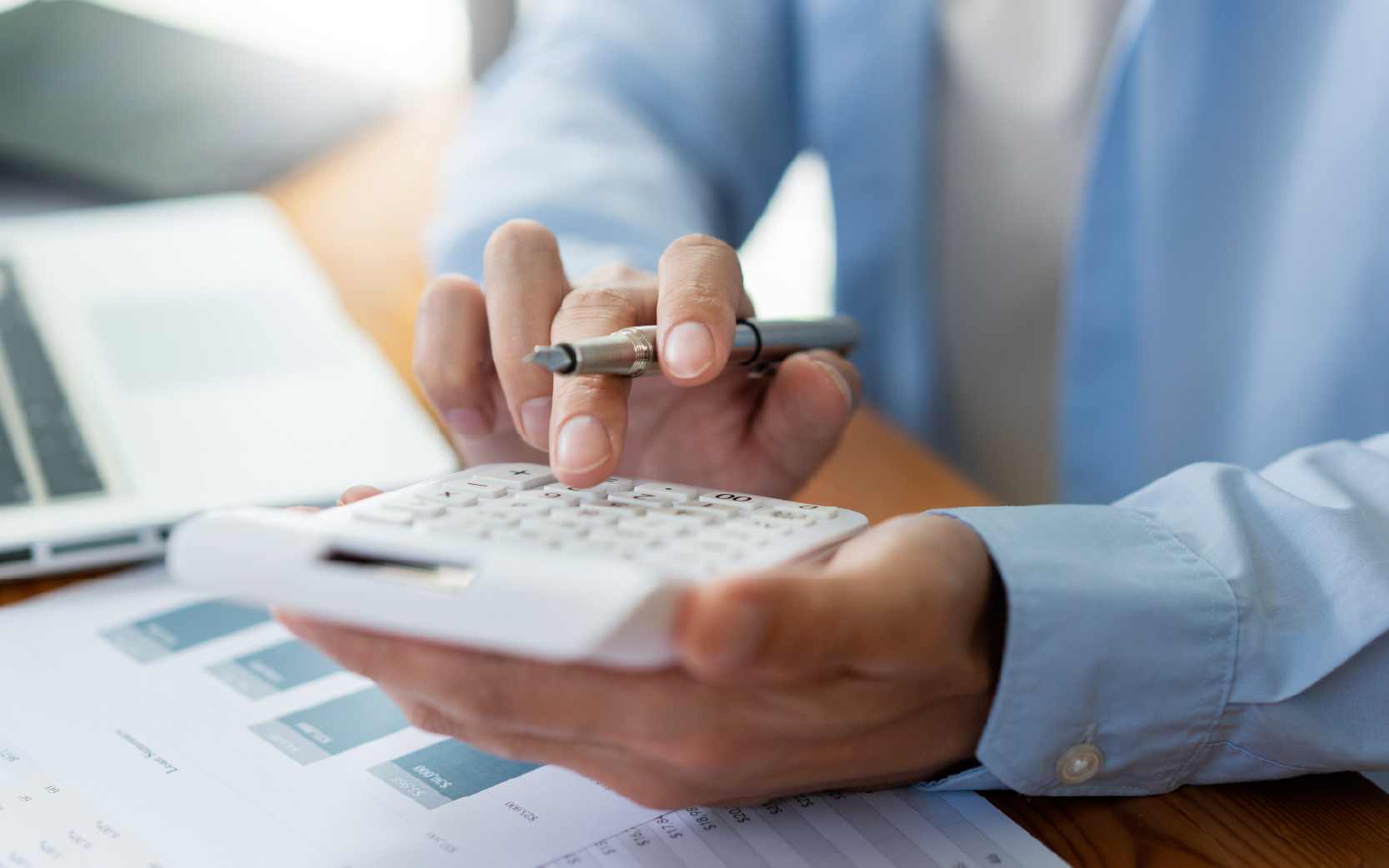 analyst using hand calculator over printed documents and laptop