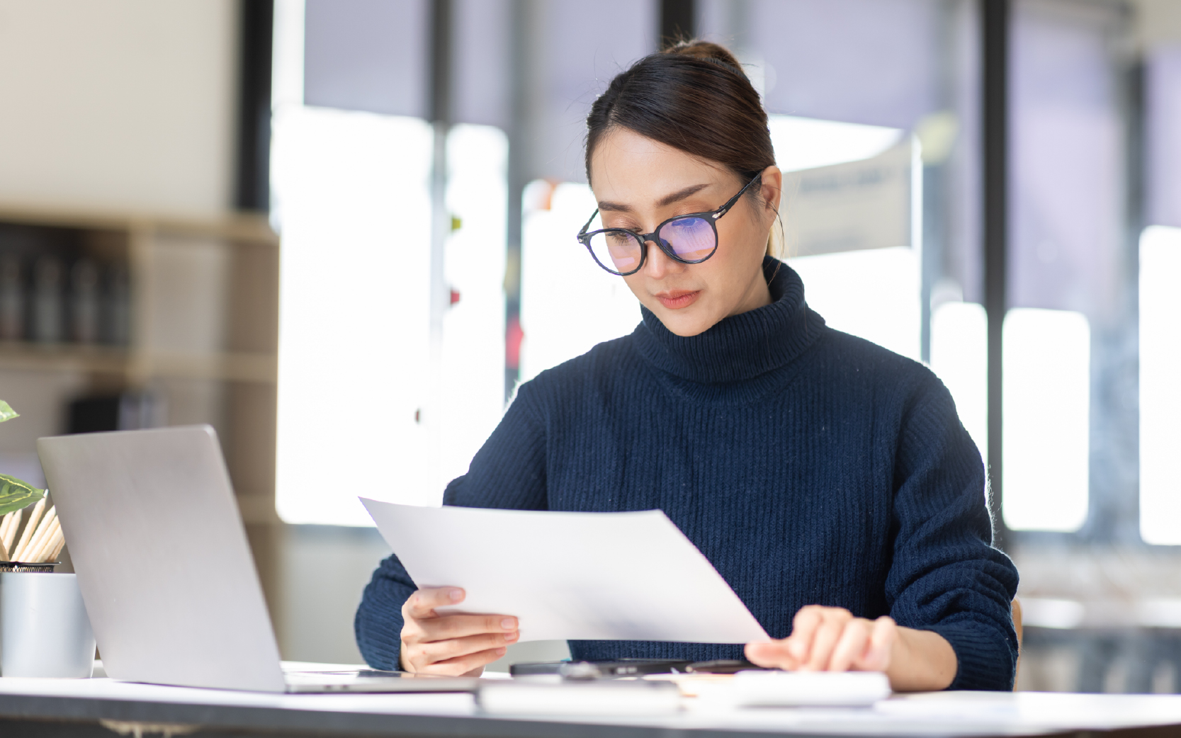 professional business woman office worker sitting at desk working on laptop reading a document and crunching numbers on a calculator